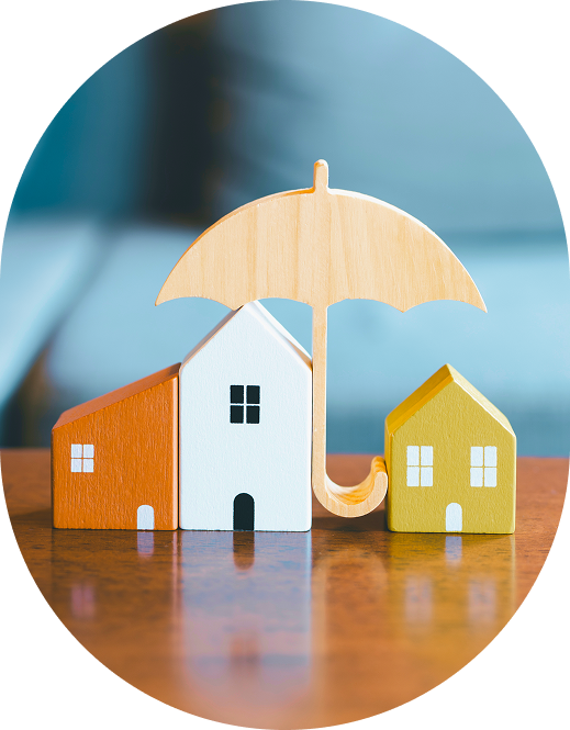 Three wooden house models of different sizes, one orange, one white, and one yellow, are arranged under a wooden umbrella on a wooden surface.
